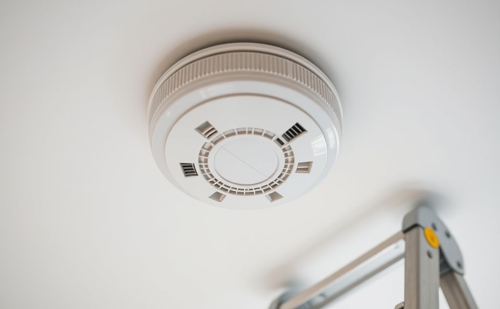 A close-up of a smoke detector mounted on a ceiling with a ladder nearby.
