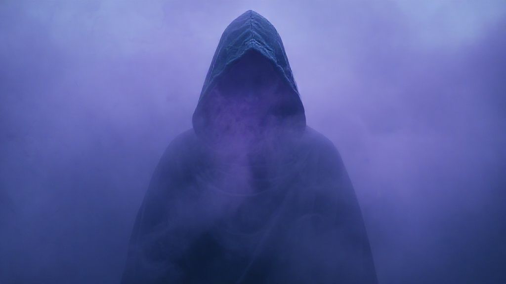 A hooded figure, draped in deep indigo, stands amidst swirling, lavender fog. Soft light emanates from within the hood, illuminating a peaceful aura. Mystic and ethereal, 4k cinematic.