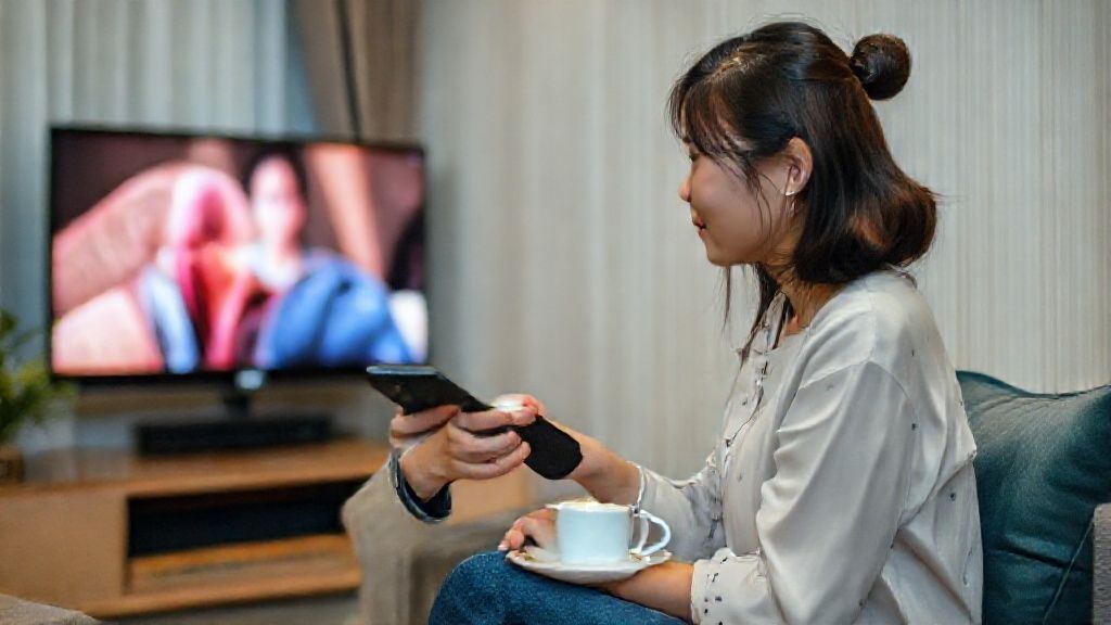Seorang wanita muda tersenyum puas sambil memegang remote TV di depan layar TV LG yang menampilkan gambar jernih. Ada cangkir kopi di meja sampingnya.