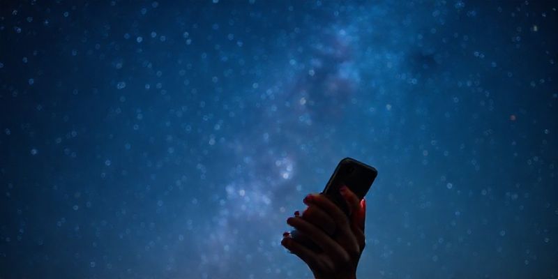 Telefonunda burcunu okuyan bir kadın ve arka planda parıldayan yıldızlı gökyüzü